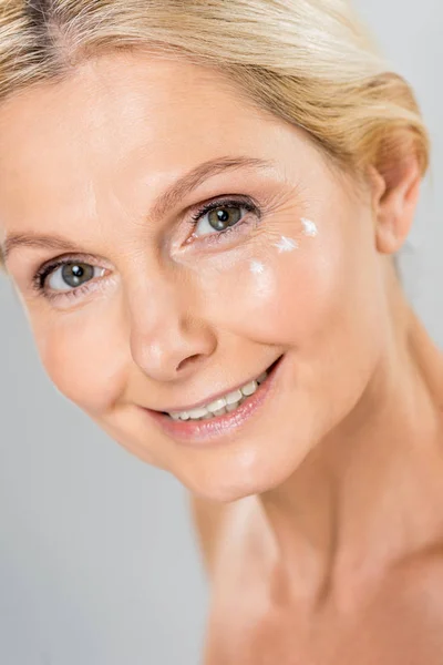 Retrato de mulher bonita e madura olhando para a câmera com creme cosmético no rosto isolado em cinza — Fotografia de Stock