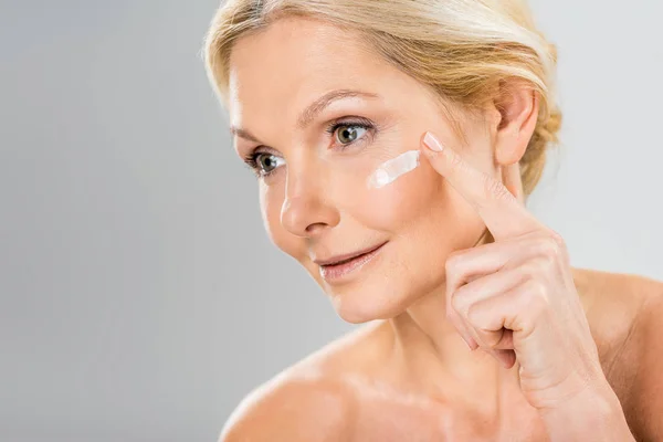Mujer hermosa y madura mirando hacia otro lado y la aplicación de crema cosmética en la cara - foto de stock
