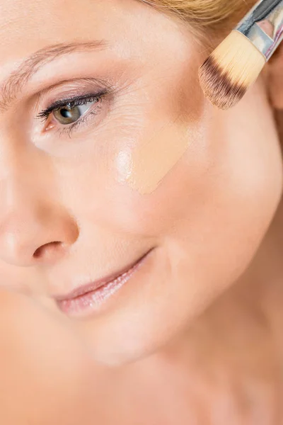 Selektiver Fokus der schönen und reifen Frau, die wegschaut und Make-up-Foundation mit kosmetischem Pinsel aufträgt — Stockfoto