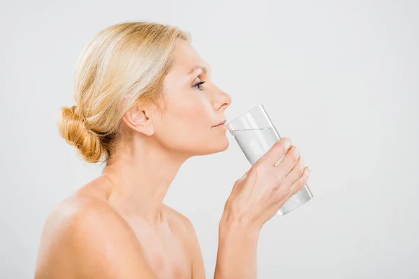 Vista lateral de la mujer madura bebiendo agua de vidrio aislado en gris - foto de stock