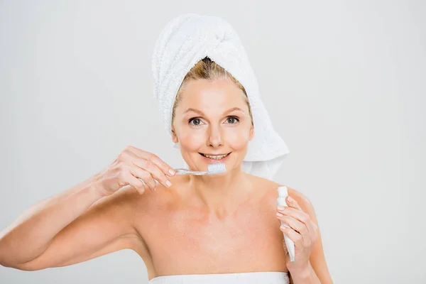 Bela e sorridente mulher madura em toalha segurando pasta de dentes e escovando dentes — Fotografia de Stock