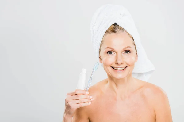 Hermosa y sonriente mujer madura en toalla sosteniendo pasta de dientes y cepillo de dientes - foto de stock