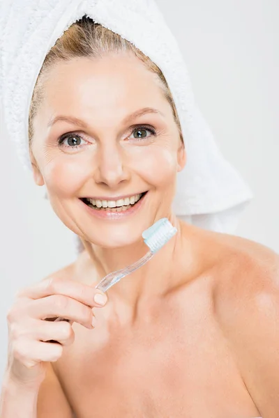 Beautiful and smiling mature woman in towel brushing teeth isolated on grey — Stock Photo