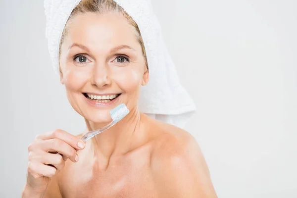 Atractiva y sonriente mujer madura en toalla cepillarse los dientes sobre fondo gris - foto de stock