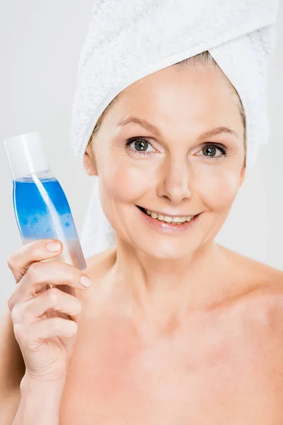 Atractiva y sonriente mujer madura en botella de toalla con agua micelar aislada en gris - foto de stock