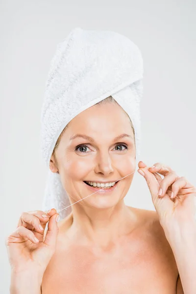 Atractiva y sonriente mujer madura en toalla cepillarse los dientes con hilo dental y mirando a la cámara - foto de stock