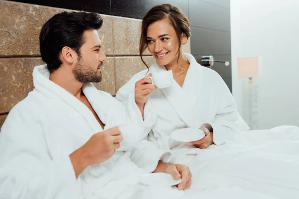 Barbudo hombre y bonita novia sosteniendo tazas con café en la cama - foto de stock