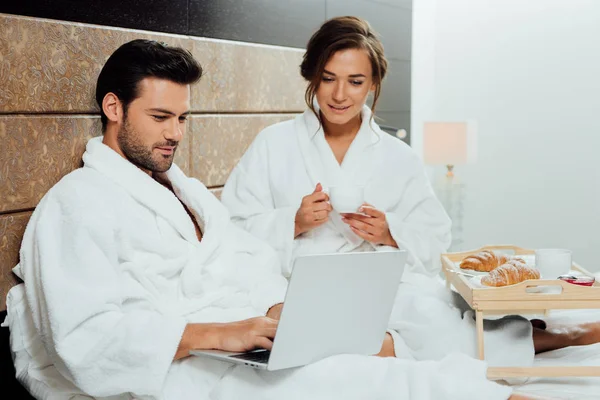 Mulher atraente segurando copo perto da bandeja com café da manhã e sentado perto do namorado olhando para laptop — Fotografia de Stock
