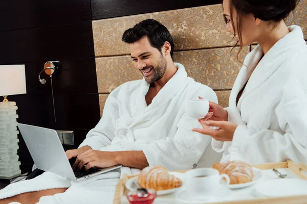 Hombre guapo usando el ordenador portátil en la cama cerca de hermosa novia con taza de café - foto de stock