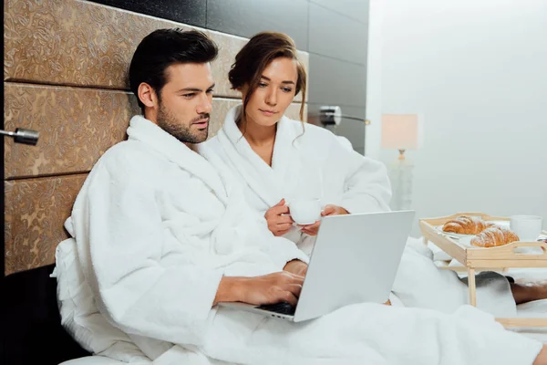 Bela mulher segurando copo e olhando para laptop com homem bonito na cama — Fotografia de Stock