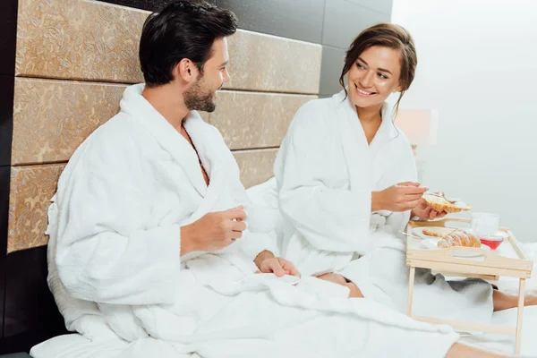 Красива жінка тримає круасан і дивиться на красивого бородатого чоловіка з чашкою кави в ліжку — стокове фото