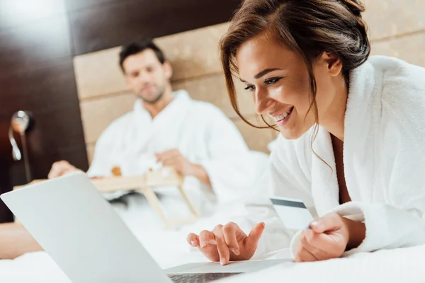 Foco seletivo da mulher alegre usando laptop enquanto segurando cartão de crédito perto do namorado — Fotografia de Stock