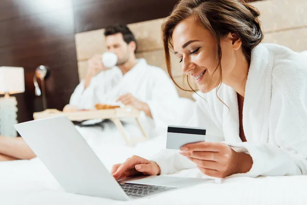 Selective focus of happy woman using laptop while holding credit card near boyfriend — Stock Photo