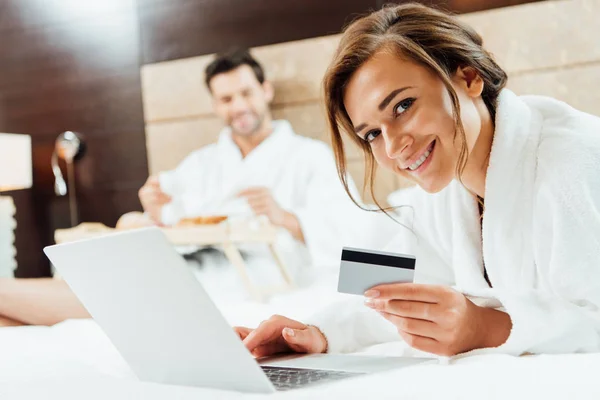 Enfoque selectivo de la mujer atractiva usando el ordenador portátil mientras sostiene la tarjeta de crédito cerca de novio - foto de stock