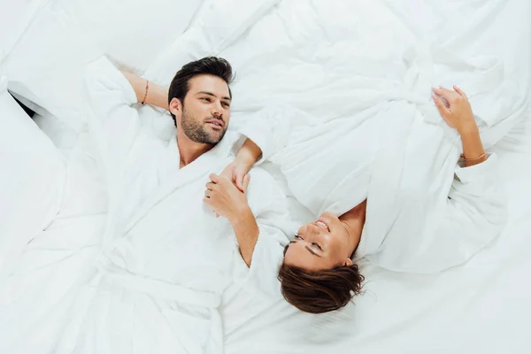 Vista superior de casal feliz em roupões de banho brancos de mãos dadas enquanto deitado na cama — Fotografia de Stock