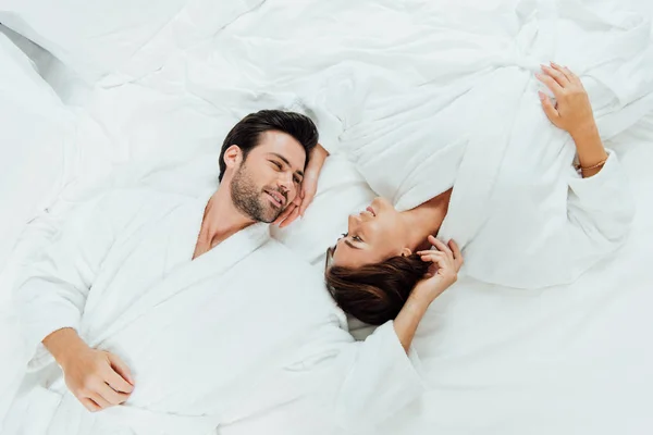 Vista superior do casal alegre em roupões de banho olhando um para o outro enquanto deitado na cama — Fotografia de Stock