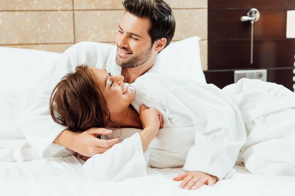 Alegre hombre en albornoz abrazando feliz novia mientras está acostado en la cama - foto de stock