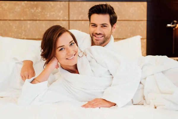 Cheerful man and happy girlfriend in white bathrobes looking at camera while lying on bed — Stock Photo