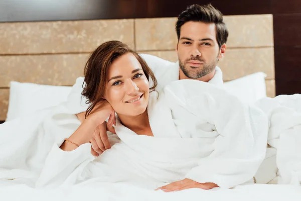 Schöner Mann und glückliche Freundin in weißen Bademänteln, die auf dem Bett liegend in die Kamera schauen — Stockfoto
