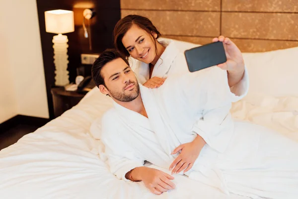 Счастливая пара делает селфи на смартфоне, лежа на кровати — стоковое фото