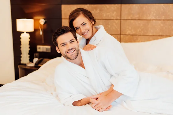 Hombre guapo y novia feliz en batas blancas abrazando y mirando a la cámara mientras está acostado en la cama - foto de stock