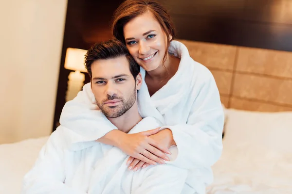Happy girlfriend in white bathrobe hugging handsome boyfriend and looking at camera — Stock Photo