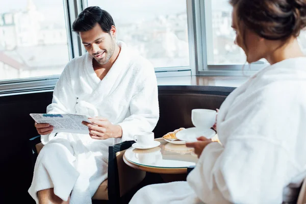 Selective focus of handsome man reading newspaper in bathrobe near woman holding cup — Stock Photo