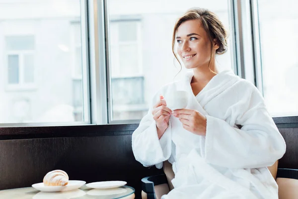 Atractiva mujer morena en albornoz sonriendo y sosteniendo la taza de café - foto de stock