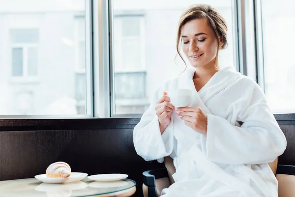 Attraktive brünette Frau im Bademantel, die lächelt und auf eine Tasse Kaffee blickt — Stockfoto