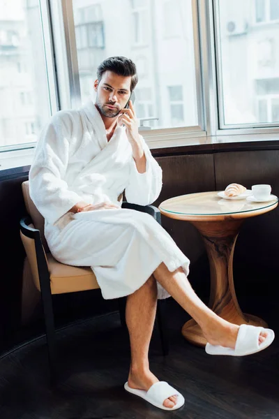 Красивий бородатий чоловік у білому халаті сидить на стільці і розмовляє на смартфоні — стокове фото