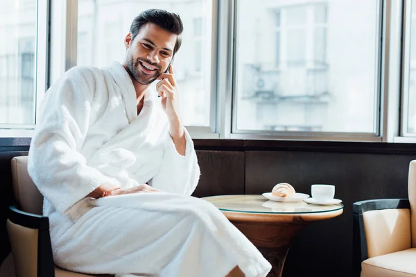 Cheerful bearded man in white bathrobe sitting on chair and talking on smartphone — Stock Photo