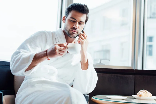 Bearded man in white bathrobe holding cup and talking on smartphone — Stock Photo