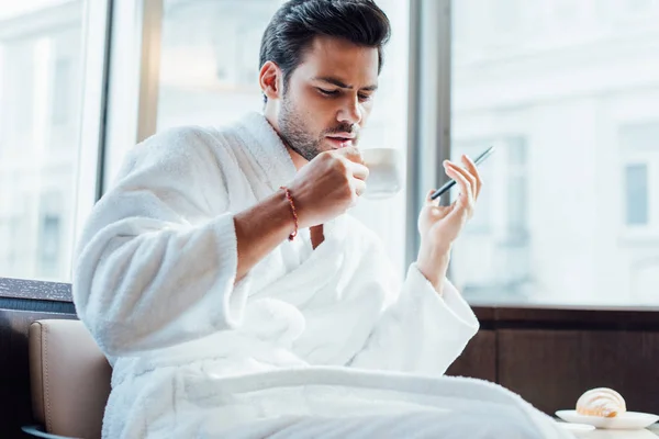 Hombre barbudo guapo en albornoz blanco sosteniendo teléfono inteligente y taza con café - foto de stock