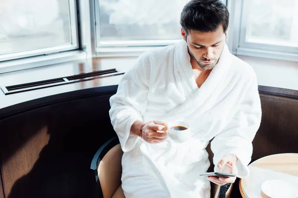 Vista aérea del hombre barbudo guapo en albornoz blanco sosteniendo teléfono inteligente y taza con café - foto de stock