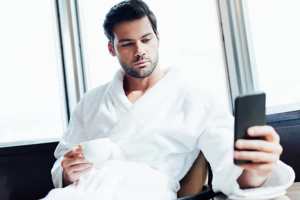 Selective focus of handsome bearded man in white bathrobe using smartphone and holding cup with coffee — Stock Photo