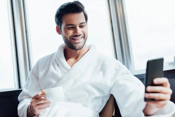 Selective focus of cheerful bearded man in white bathrobe using smartphone and holding cup with coffee — Stock Photo