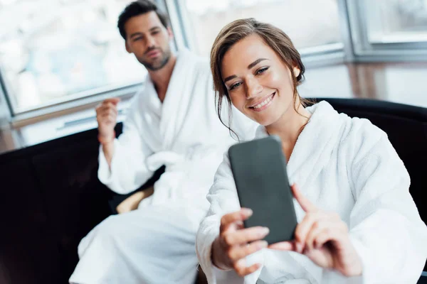 Selective focus of cheerful woman in bathrobe taking selfie with handsome boyfriend in hotel — Stock Photo