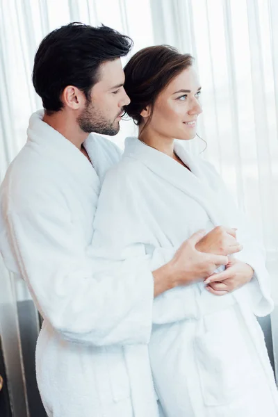 Feliz pareja en batas blancas abrazándose en el hotel - foto de stock