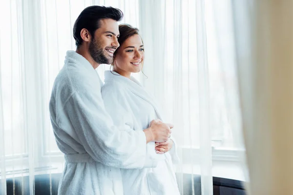 Enfoque selectivo de hombre alegre abrazando a mujer feliz en albornoz blanco en el hotel - foto de stock