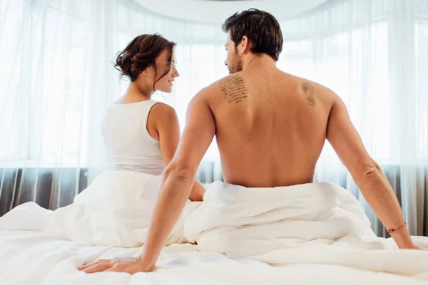 Attractive brunette woman looking at tattooed boyfriend while sitting on bed — Stock Photo