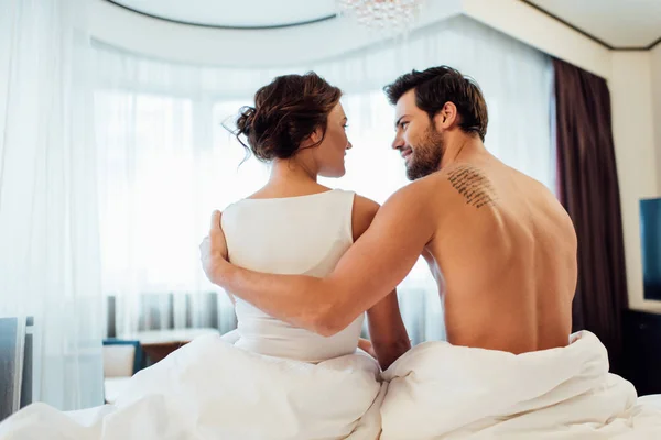 Tattooed boyfriend hugging attractive brunette woman while sitting on bed — Stock Photo