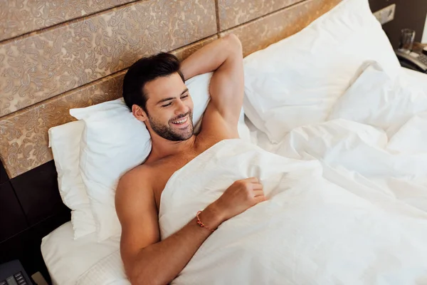 Blick von oben auf einen fröhlichen bärtigen Mann, der unter einer Decke im Bett liegt — Stockfoto