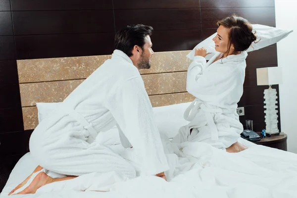 Happy couple in white bathrobes having pillow fight on bed — Stock Photo