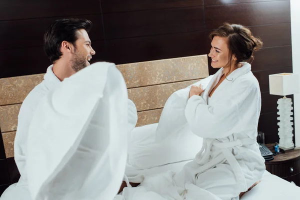 Alegre pareja en albornoces teniendo almohada lucha en cama en hotel - foto de stock
