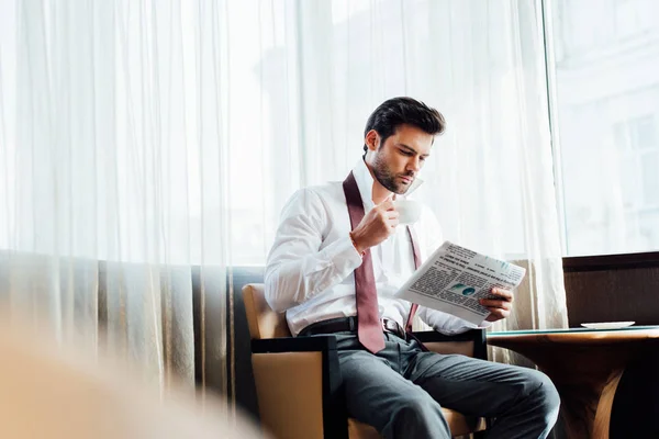 Избирательный фокус красивого мужчины в костюме, сидящего возле кофейного столика, читающего газету и держащего чашку кофе — стоковое фото