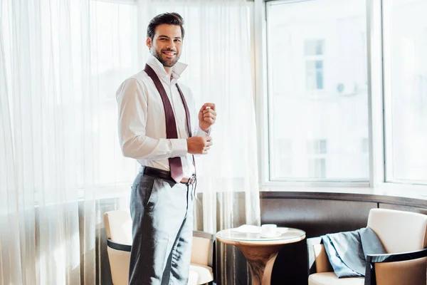 Веселий бородатий чоловік у костюмі, що торкається сорочки, стоячи в готелі — стокове фото