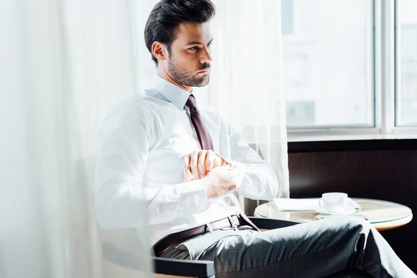 Handsome bearded man in suit touching shirt while sitting near coffee table with cup of coffee — Stock Photo
