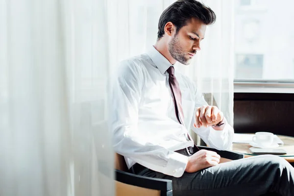 Selektiver Fokus des bärtigen Geschäftsmannes im Anzug, der auf die Uhr schaut, während er am Kaffeetisch sitzt — Stockfoto