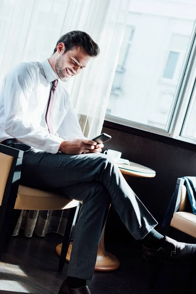 Cheerful businessman in suit looking at smartphone while sitting near coffee table in hotel — Stock Photo