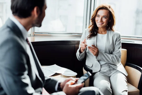 Selective focus of cheerful businesswoman holding cup and looking at businessman with smartphone — Stock Photo